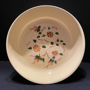 May include: A cream-colored ceramic bowl with a floral design. The interior features white flowers with pink accents and green leaves. The bowl has a wide rim and a smooth, glossy finish.