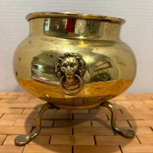 May include: A vintage brass pot with a lion's head ring handle and three curved legs. The pot has a rounded body and a wide rim. The brass has a polished, reflective surface.