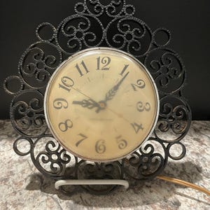 May include: A black metal clock with an ornate frame and a white face. The clock has black hands and black numbers. The clock is plugged in and has a cord.