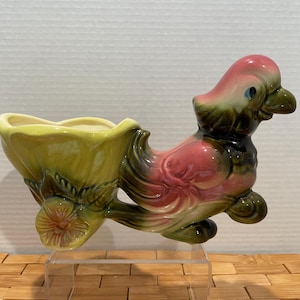 May include: A vintage ceramic planter in the shape of a bird. The bird has a pink head and chest, with green and black accents. The planter section is yellow and green, with a floral design. A decorative piece.