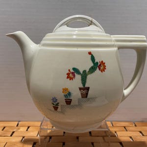 May include: Cream-colored ceramic teapot with a lid and handle. The teapot features a painted design of cacti in pots with orange, yellow, and blue flowers. The teapot is displayed on a clear stand.