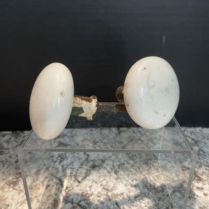May include: Two antique white porcelain door knobs with a mottled appearance. The knobs are oval-shaped and mounted on a tarnished metal plate. The background is a dark color.