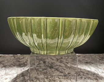 May include: A green ceramic bowl with a ribbed exterior. The bowl has a wide, oval shape and sits on a clear, rectangular stand. The bowl's color is a muted, earthy green.