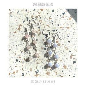May include: A pair of dangle earrings with silver chains and crystal beads. One earring has light pink beads and the other has light blue beads. The earrings are on a light gray and brown speckled surface. "DANGLY CRYSTAL EARRINGS" and "ROSE QUARTZ + BLUE LACE AGATE" are written in white text below the earrings.