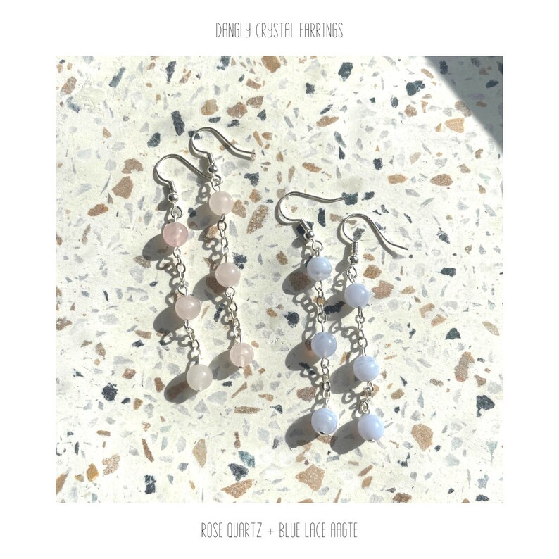 May include: A pair of dangle earrings with silver chains and crystal beads. One earring has light pink beads and the other has light blue beads. The earrings are on a light gray and brown speckled surface. "DANGLY CRYSTAL EARRINGS" and "ROSE QUARTZ + BLUE LACE AGATE" are written in white text below the earrings.