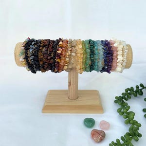 Peut inclure: Présentation de bracelets de pépites de pierres précieuses colorées sur un présentoir en bois. Les bracelets sont disposés en arc-en-ciel, avec des couleurs comme le noir, le marron, l'orange, le jaune, le vert, le bleu, le violet et le blanc. Trois pierres précieuses en forme de cœur sont au premier plan.