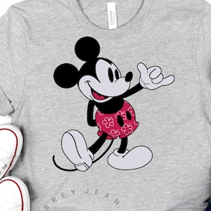 May include: A gray t-shirt with a black and white cartoon image of Mickey Mouse giving a shaka sign. The text "Aubrey Jean SVG" is printed in a circle around the image.