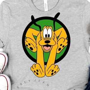 以下が含まれることがあります： 緑色の円の中にプルートの漫画の画像が描かれた灰色のTシャツ。Tシャツには「Aubrey Jean SVG」という文字が印刷されています。