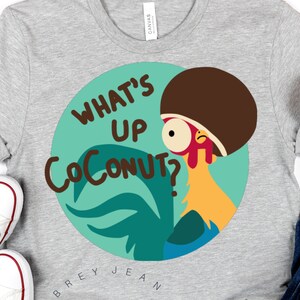 Puede incluir: Una camiseta gris con un gallo de dibujos animados que lleva un afro marrón y el texto "What's Up Coconut?" impreso en ella. La camiseta está sobre una superficie blanca con un par de zapatillas blancas y un par de vaqueros azules con una diadema de purpurina dorada.