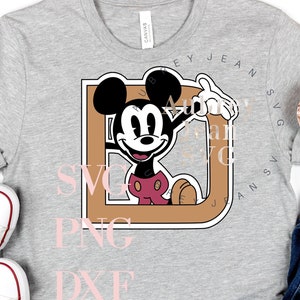 May include: A gray t-shirt with a brown and black Mickey Mouse character in a brown frame. The text "SVG PNG DXF" is printed in pink below the frame. The text "KEY JEAN SVG" is printed in black around the frame.