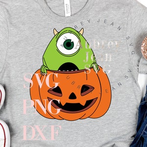 May include: A heather gray t-shirt featuring a green one-eyed monster graphic sitting inside an orange jack-o'-lantern pumpkin. The shirt includes the text SVG, PNG, DXF, and Aubrey Jean SVG. White canvas sneakers and blue denim jeans are also visible.