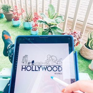 May include: A digital drawing of a Hollywood Studios logo on a tablet screen. The logo is a black and white illustration of a cityscape with the words "HOLLYWOOD STUDIOS" underneath.