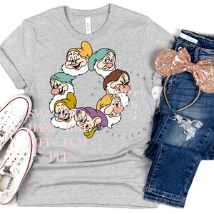 K&ouml;nnte beinhalten: Ein graues T-Shirt mit einem Kreis aus sieben Zwergen aus dem Disney-Film Schneewittchen. Die Zwerge sind in einem Kreis angeordnet, mit ihren Namen darum herum. Das T-Shirt liegt auf einem wei&szlig;en Hintergrund mit einem Paar wei&szlig;en Turnschuhen, einer blauen Jeans und einem Paar goldenen Glitzer-Minnie-Maus-Ohren.