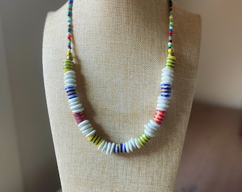 Puede incluir: Un collar de cuentas de colores con cuentas blancas, amarillas, rojas, azules y verdes. El collar está en un maniquí beige.