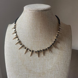May include: A gold and black beaded necklace with gold spikes. The necklace is on a mannequin.