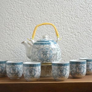 Peut inclure: Service à thé en céramique avec une théière et six tasses assorties. La théière a une anse jaune. L'ensemble présente un motif floral blanc et bleu et un caractère chinois. L'ensemble est présenté sur une surface en bois.