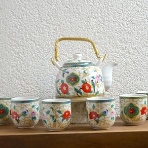 Peut inclure: Service à thé en céramique blanche avec un motif floral, comprenant une théière et six tasses assorties. La théière a une anse et un bec dorés. Les tasses et la théière sont décorées de fleurs et d'oiseaux rouges, roses et bleus.