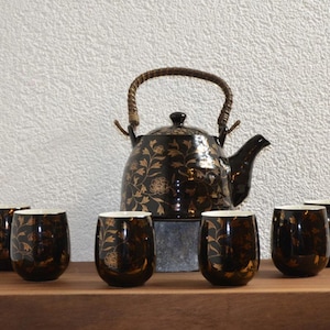 Peut inclure: Service à thé noir et doré comprenant une théière et six tasses assorties. La théière a une anse tressée et un motif floral. Les tasses et la théière ont une finition brillante, posées sur une surface en bois.