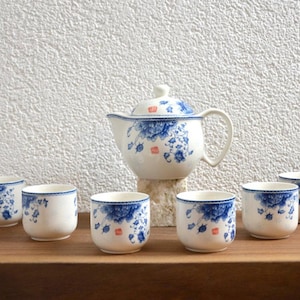 Op de afbeelding: Een witte keramische theeset met blauwe bloemmotieven en rode accenten. De set bevat een theepot en zes bijpassende kopjes. De theepot staat op een kleine steen en de kopjes zijn op een houten oppervlak geplaatst.