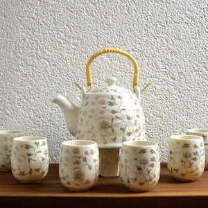 Peut inclure: Service à thé en céramique blanche avec un motif floral dans des tons or et marron. L'ensemble comprend une théière avec une anse jaune et six tasses assorties. L'ensemble est présenté sur une surface en bois.