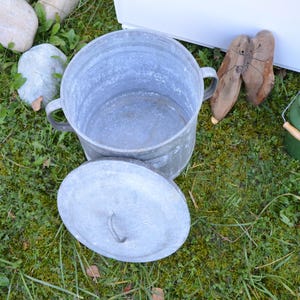 Op de afbeelding: Een gegalvaniseerde metalen emmer met deksel en handvatten, en een groene melkkan met houten handvat staan op gras. Twee bruine leren schoenen zijn ook zichtbaar. De scène speelt zich buiten af.