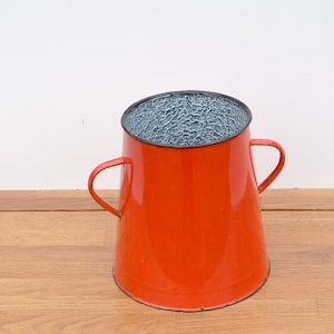 May include: A vibrant, red enamelware pitcher with two handles. The interior has a textured, gray surface. The pitcher sits on a wooden surface, suggesting a rustic or vintage aesthetic. The bright color and design make it a decorative kitchen item.
