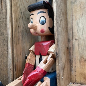 Könnte beinhalten: Eine Holzpuppe mit langer Nase, schwarzen Haaren und bemalten Gesichtszügen. Die Figur trägt ein rotes Hemd, eine blaue Hose und rote Handschuhe, mit durch Gelenke verbundenen Gliedmaßen. Die Puppe befindet sich in einer hölzernen Kistenstruktur.