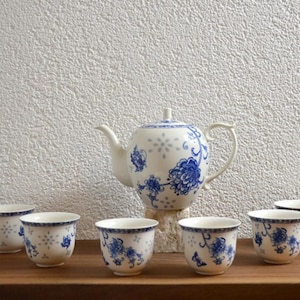 Peut inclure: Service à thé en porcelaine blanche avec motifs floraux bleus. L'ensemble comprend une théière et six petites tasses. La théière a une anse et un bec courbés. Les tasses et la théière sont disposées sur une surface en bois.