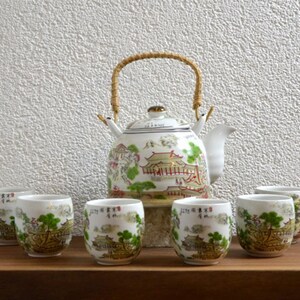 Peut inclure: Service à thé en céramique blanche comprenant une théière avec une anse tressée et six tasses assorties. L'ensemble est décoré d'un paysage asiatique traditionnel en vert, rouge et or. La théière a un dessus doré.