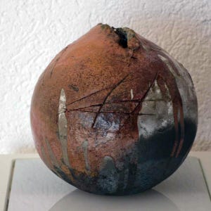 Könnte beinhalten: Eine handgefertigte, kugelförmige Keramikvase mit strukturierter Oberfläche. Die Vase hat einen Farbverlauf von rötlich-braun zu dunkelblau, mit silberfarbenen Akzenten und geätzten Linien. Die Oberseite hat eine kleine Öffnung.