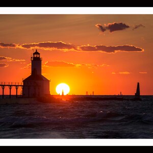 May include: Silhouette of a lighthouse on a pier with the sun setting over the water. The sky is a vibrant orange and red, with clouds in the distance. The city skyline is visible in the background.
