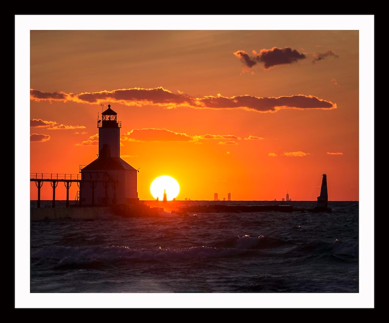 May include: Silhouette of a lighthouse on a pier with the sun setting over the water. The sky is a vibrant orange and red, with clouds in the distance. The city skyline is visible in the background.