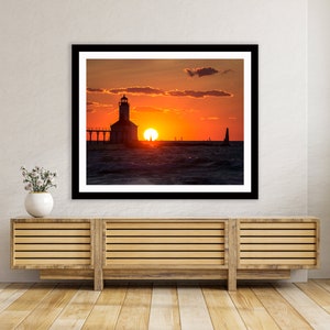 May include: A black and white photograph of a lighthouse silhouetted against a vibrant orange and red sunset over a body of water. The lighthouse is on a pier with a few boats in the distance.