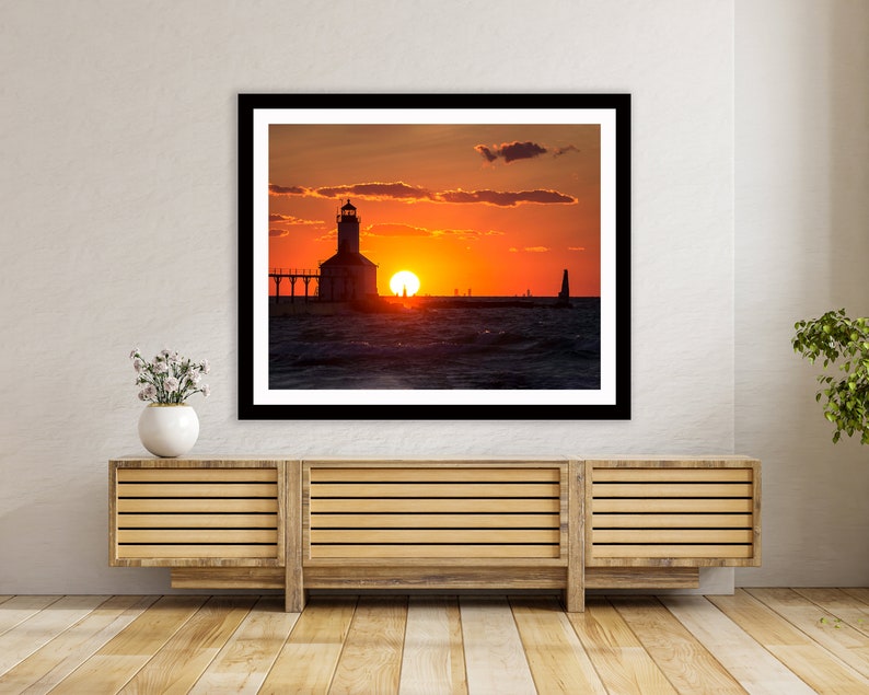 May include: A black and white photograph of a lighthouse silhouetted against a vibrant orange and red sunset over a body of water. The lighthouse is on a pier with a few boats in the distance.