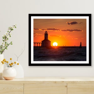 May include: A silhouette of a lighthouse on a pier with the sun setting over the water. The sky is a vibrant orange and red, with clouds in the background.