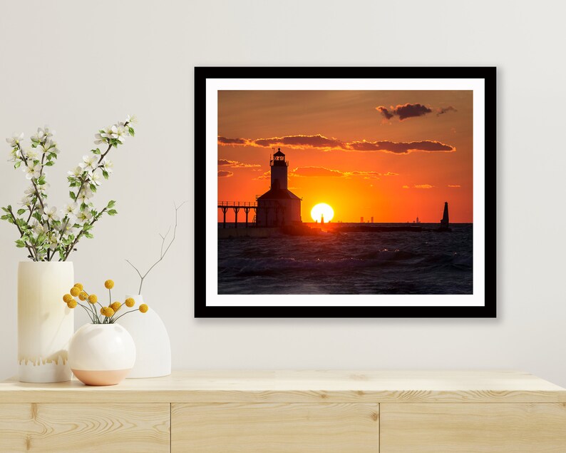 May include: A silhouette of a lighthouse on a pier with the sun setting over the water. The sky is a vibrant orange and red, with clouds in the background.