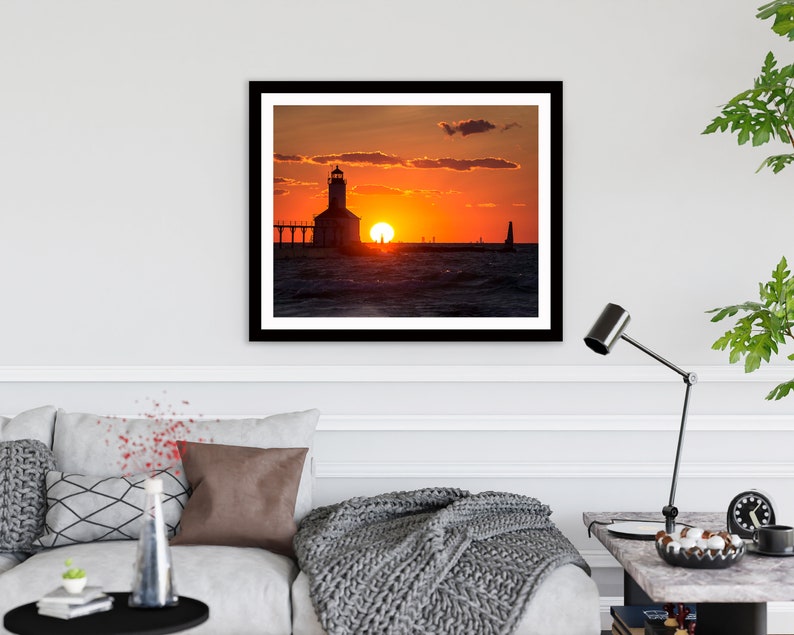 May include: A silhouette of a lighthouse on a pier with the sun setting over the water. The sky is a vibrant orange and pink.