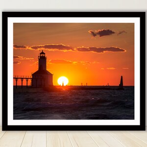 May include: A silhouette of a lighthouse on a pier with the sun setting over the water. The sky is a vibrant orange and red, with clouds in the distance.
