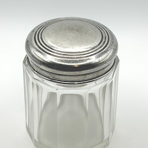 May include: A clear glass jar with a faceted design and a silver-colored lid. The lid is round with concentric rings. The jar has an octagonal base and a frosted interior. The jar is likely used for storing small items.
