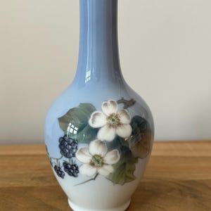 May include: A delicate, light blue porcelain vase with a white base. It features hand-painted white flowers, green leaves, and dark purple berry clusters. The vase has a narrow neck and a rounded body, perfect for displaying flowers.