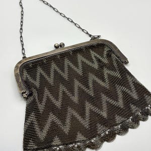 May include: Antique silver-tone metal mesh purse with a chain handle. The purse features a zigzag pattern in shades of gray and brown. The bottom edge has a scalloped trim. The clasp is a silver-tone metal with two small ball details.