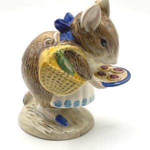 May include: A ceramic mouse figurine, adorned with a blue bow and a white dress with blue floral patterns. The mouse carries a yellow basket and a small plate with pastries. The figurine features brown and tan fur and rests on a light grey base.