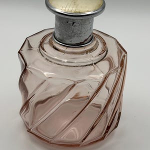 May include: A vintage pink glass perfume bottle with a silver-toned metal collar and a cream-colored top. The bottle has a unique, twisted design and is empty. The bottle is likely from the mid-20th century.
