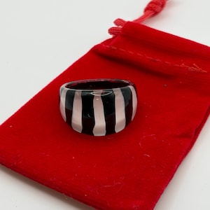 May include: A bold ring featuring a black and white striped design. The ring is displayed on a red velvet pouch with a drawstring. A striking fashion accessory.