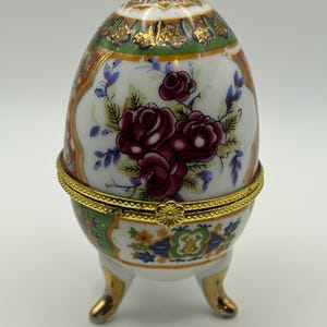 May include: A decorative porcelain egg-shaped trinket box. The egg features a floral design with burgundy roses and gold accents. It sits on a gold-trimmed base with three gold-colored feet. The top of the egg has a colorful, ornate pattern.