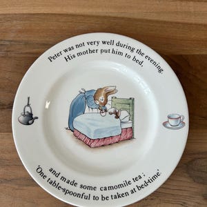 May include: A white ceramic plate with a rabbit illustration in bed, surrounded by text. The plate includes a teapot and teacup design. The text reads, "Peter was not very well during the evening. His mother put him to bed, and made some camomile tea: One table-spoonful to be taken at bed-time."