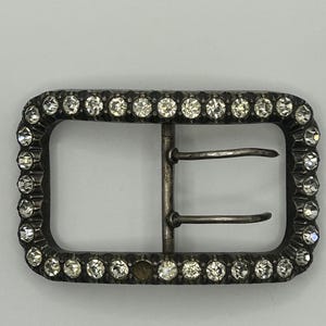 May include: Rectangular silver-toned belt buckle with a border of clear, sparkling rhinestones. The buckle has a classic design with two horizontal bars for the belt strap. The rhinestones are evenly spaced around the frame.