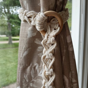 May include: A white cotton rope curtain tieback with a wooden ring. The tieback is looped around the curtain and secured with the ring.