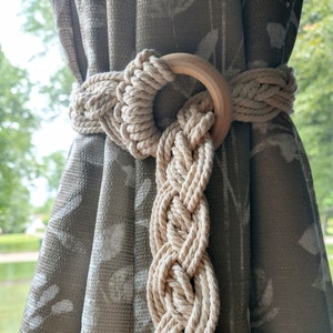 May include: A beige rope curtain tieback with a wooden ring. The tieback is braided and has a simple, minimalist design.