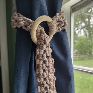 May include: A beige crocheted curtain tieback with a wooden ring. The tieback is looped around a dark blue curtain.